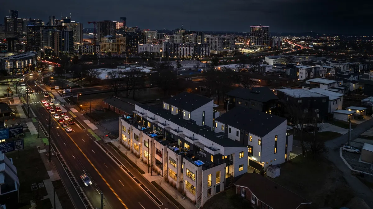 A vibrant exterior view of modern, dual townhouses set against the backdrop of the city skyline, with streetlights and vehicles adding urban energy. Rooftop decks are visible, hinting at evening gatherings and panoramic city views.