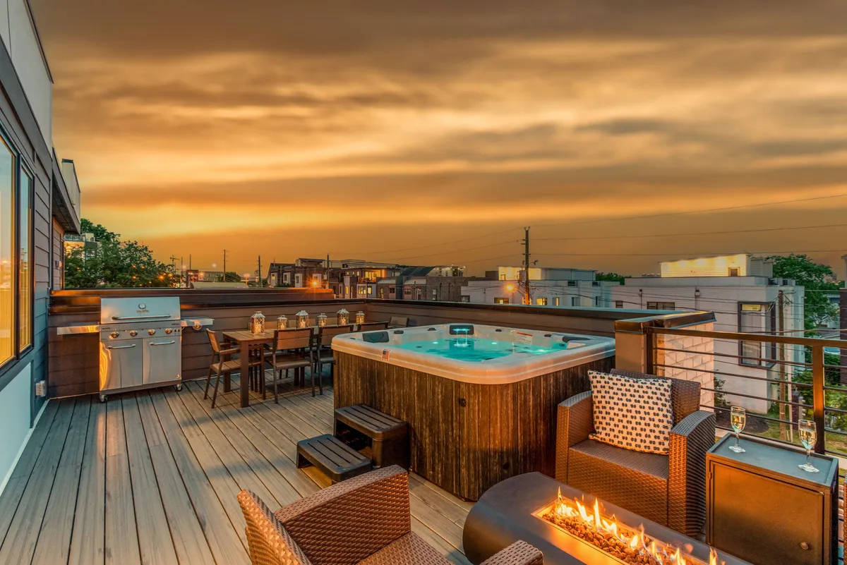 The image showcases the stylish exterior of the Edgehill Duo townhomes at sunset, with a welcoming outdoor area featuring a bubbling hot tub, a roaring fire pit surrounded by comfortable seating, a spacious dining table set for guests, and a modern grill ready for barbecues. The scene suggests a perfect spot for social gatherings and relaxation under the Nashville sky.