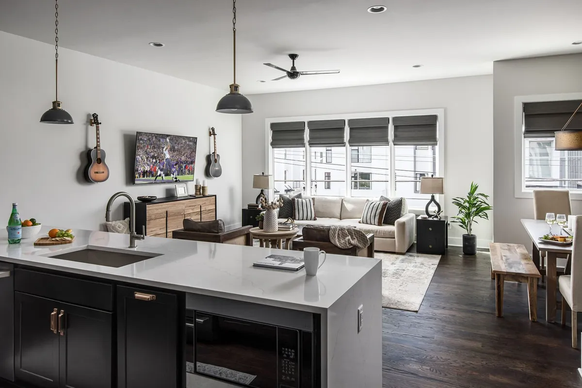 An open-concept common space featuring a kitchen island with granite countertop, a large TV displaying a sports event, a comfortable seating area with a sofa, guitars mounted on the wall, and a dining table set for two. The room blends entertainment, relaxation, and music, making it ideal for both lively group events and intimate gatherings.
