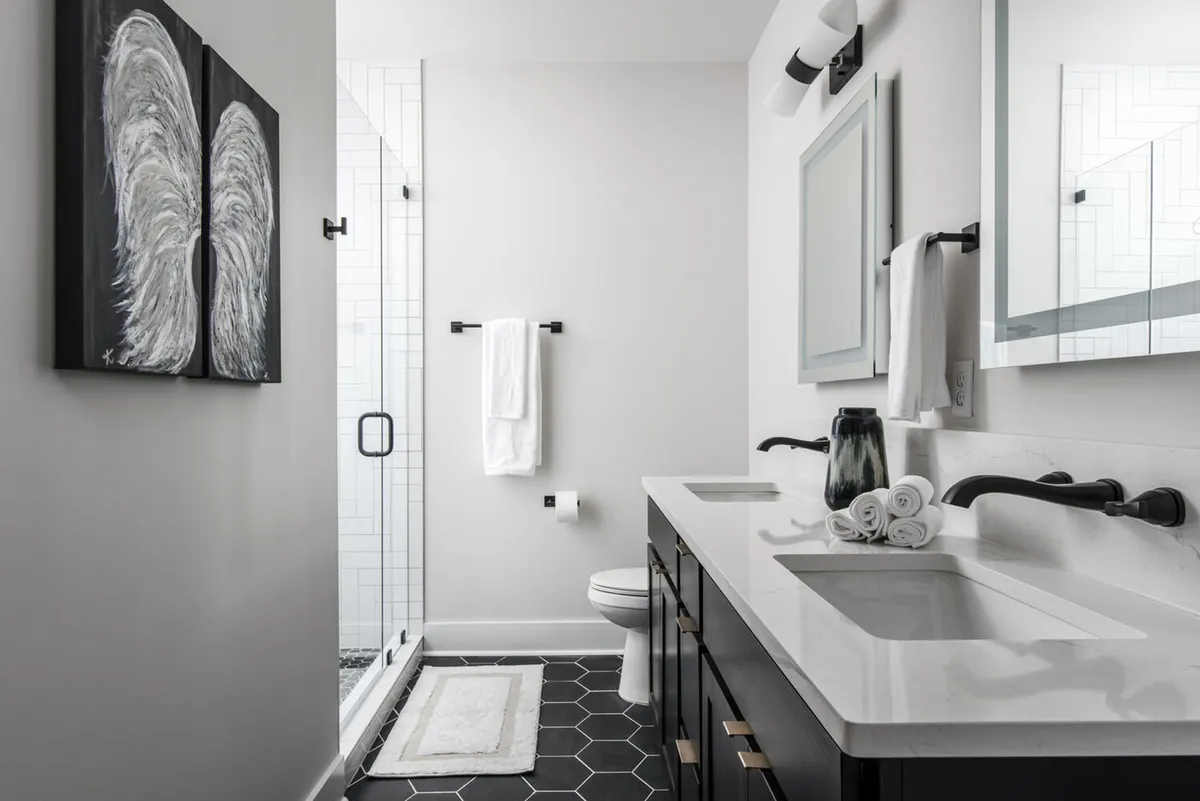 A modern bathroom with a stylish vanity featuring dual sinks, a large framed mirror, wall artwork, and a glass shower enclosure. The space is thoughtfully designed for both function and relaxation, offering a spa-like experience.