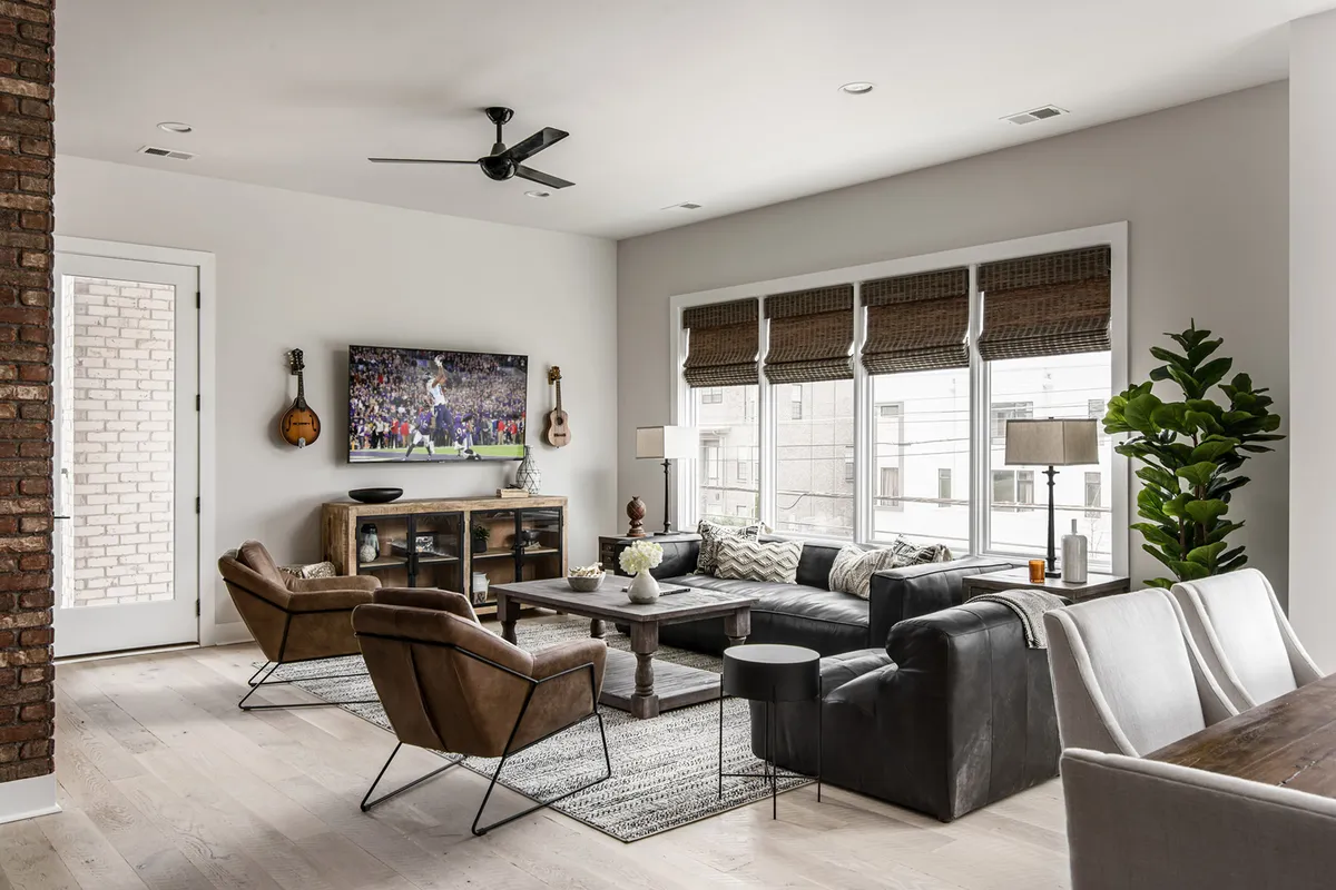 A spacious living room featuring a large sectional sofa positioned around a flat-screen television, with a wooden coffee table and window seating. The space is bathed in natural light, offering a casual, sociable atmosphere ideal for group activities like watching movies or sports and gathering with friends or family.