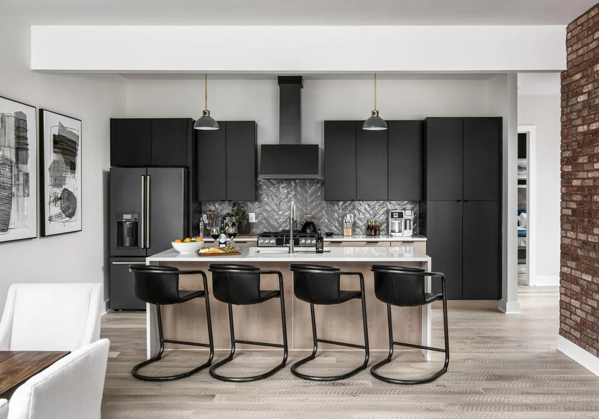 A modern kitchen and dining area with a sleek island, bar stools, black cabinetry, a chevron-patterned backsplash, and a dining table. The space is designed for cooking, entertaining, and sharing meals, exuding a contemporary and welcoming mood.