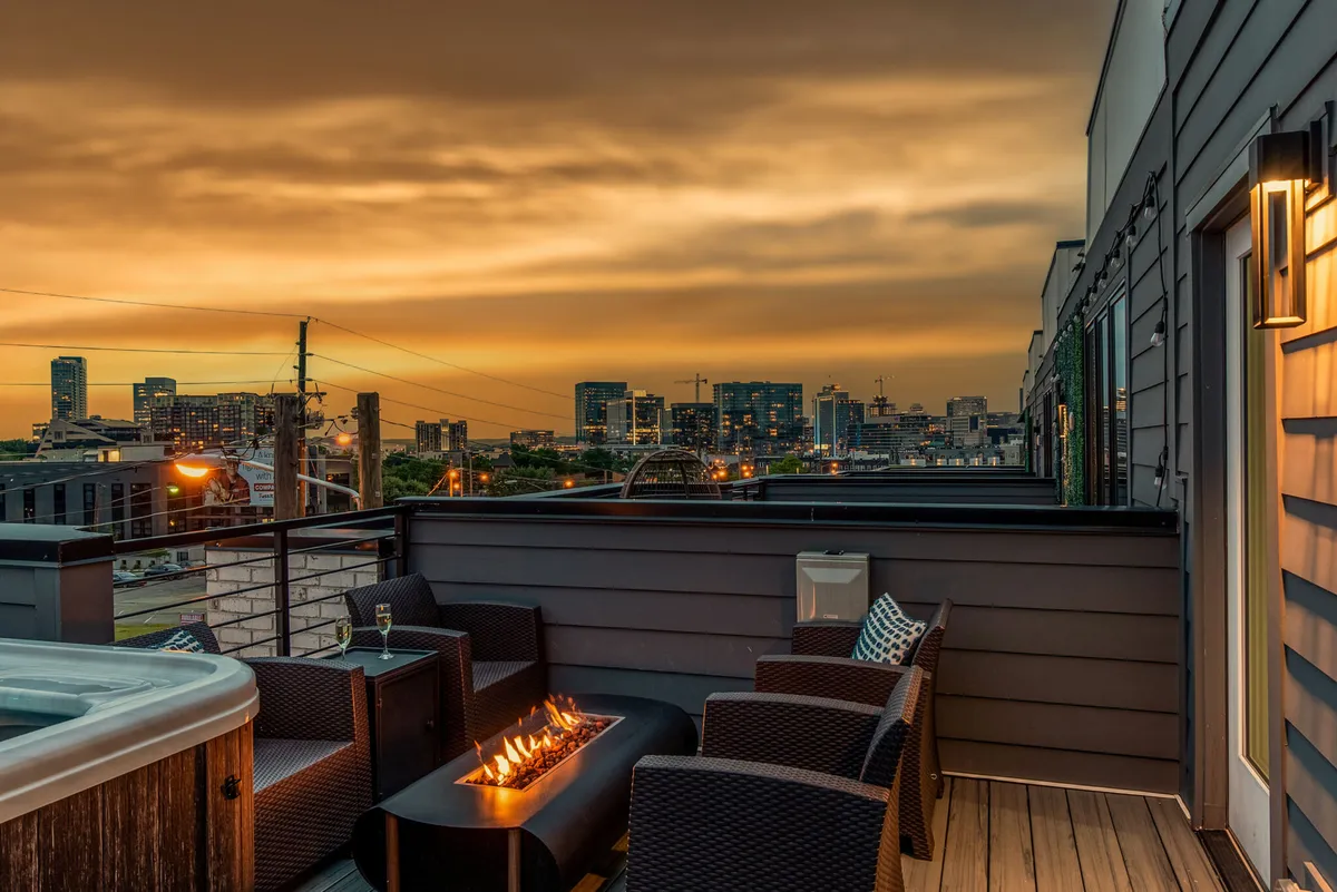 A cozy rooftop space with a fire pit surrounded by outdoor seating, all set against a sunset sky and the city skyline. The glowing fire pit creates an inviting atmosphere ideal for evening gatherings, relaxation, or stargazing above the city.