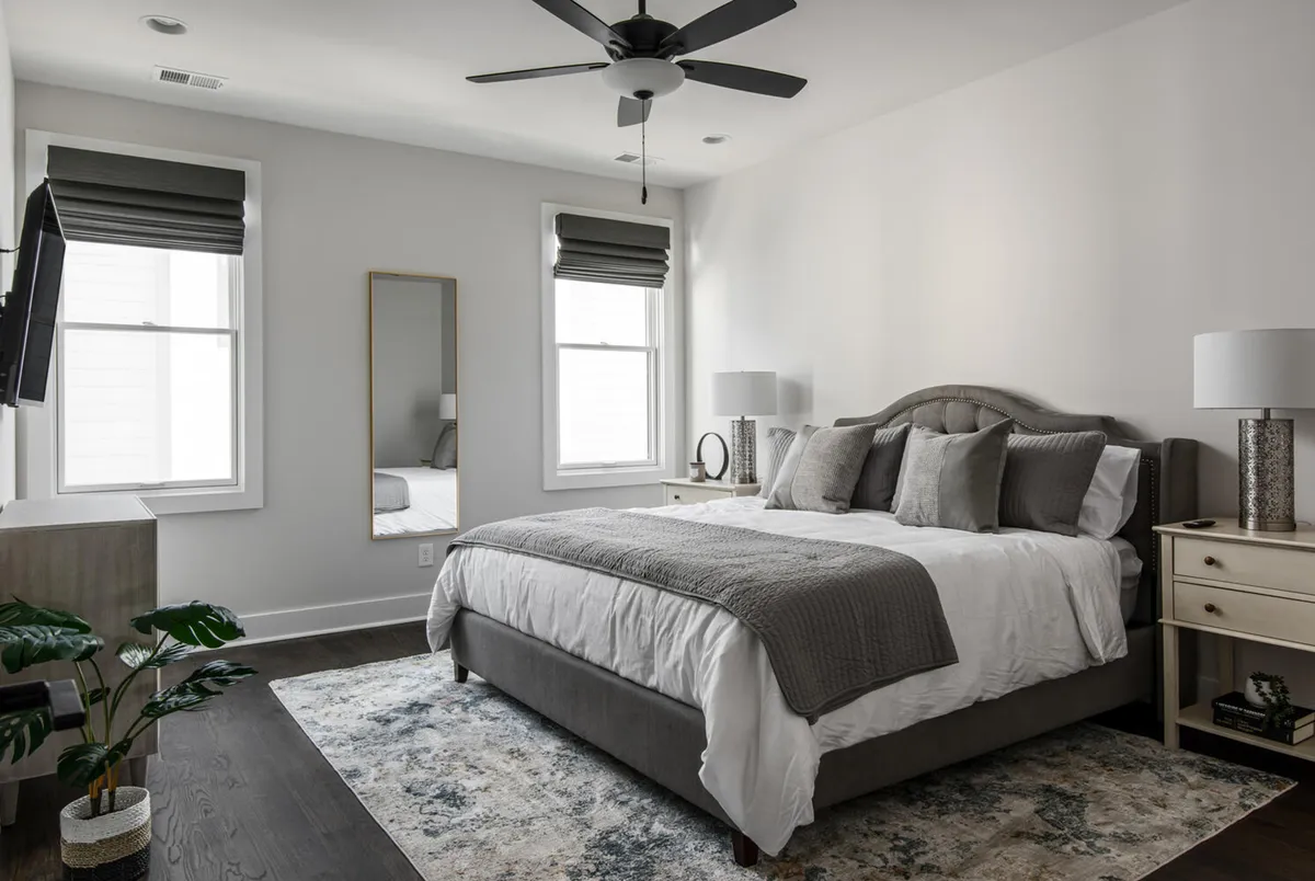 A serene bedroom featuring a plush king-size bed with grey bedding, decorative pillows, mirrored wall, and nightstands with lamps. The space is bathed in natural light, creating a tranquil atmosphere perfect for relaxation and restful sleep.